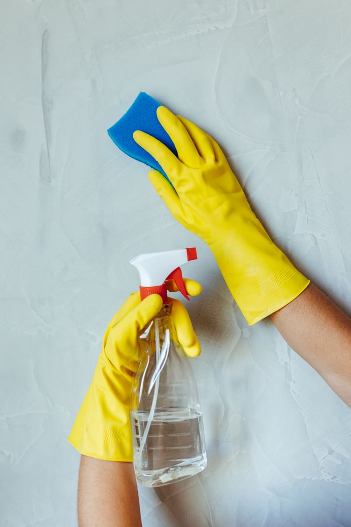 Mains gantées de jaune nettoyant un mur avec une éponge et un pulvérisateur.